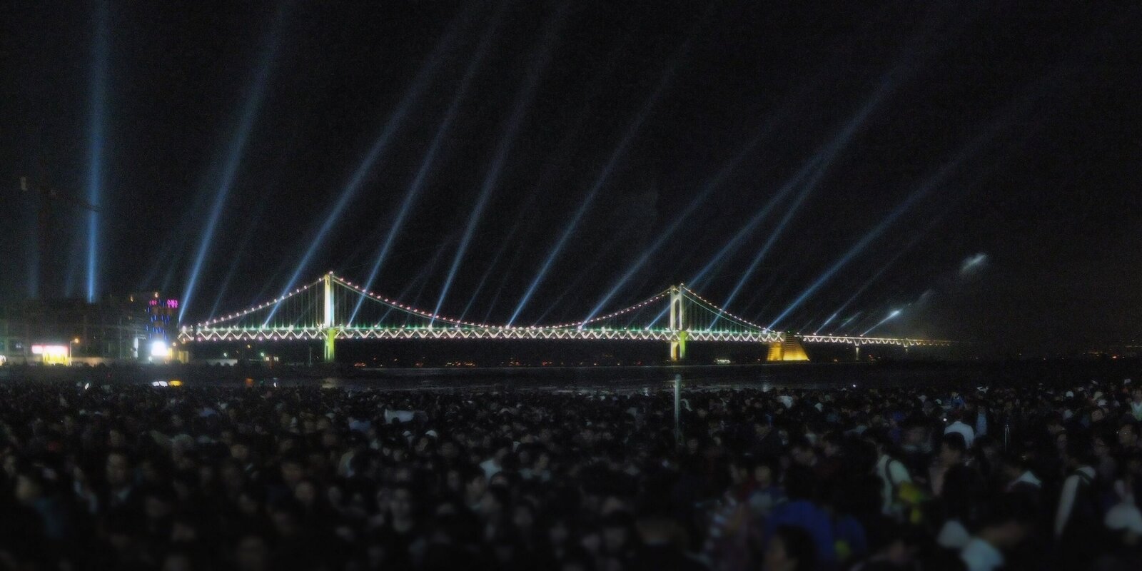 조명이 켜진 광안대교 앞 백사장을 사람들이 빈틈없이 메운 밤 풍경. 하늘로 서치라이트 불빛이 여러 줄 뻗어 있다