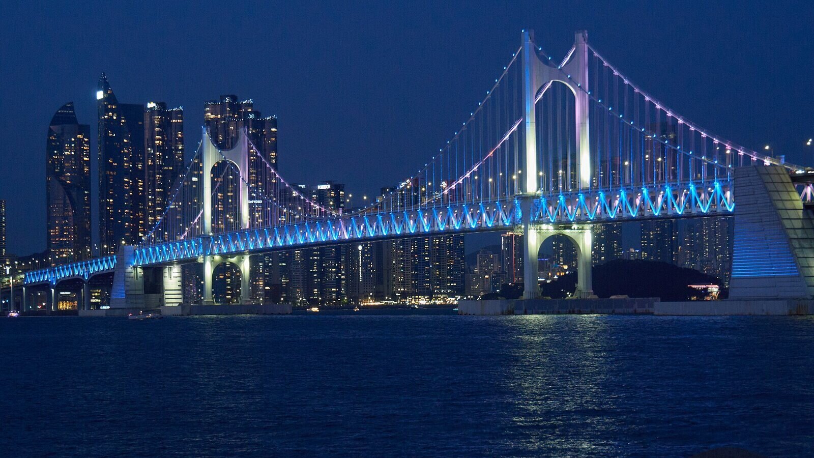 밤에 광안리 바닷가에서 가까이 본 광안대교. 다리 옆면에 파란 조명이 켜져 있고, 다리 뒤로 불 켜진 마린시티 고층 건물들과 어두운 동백섬이 보인다