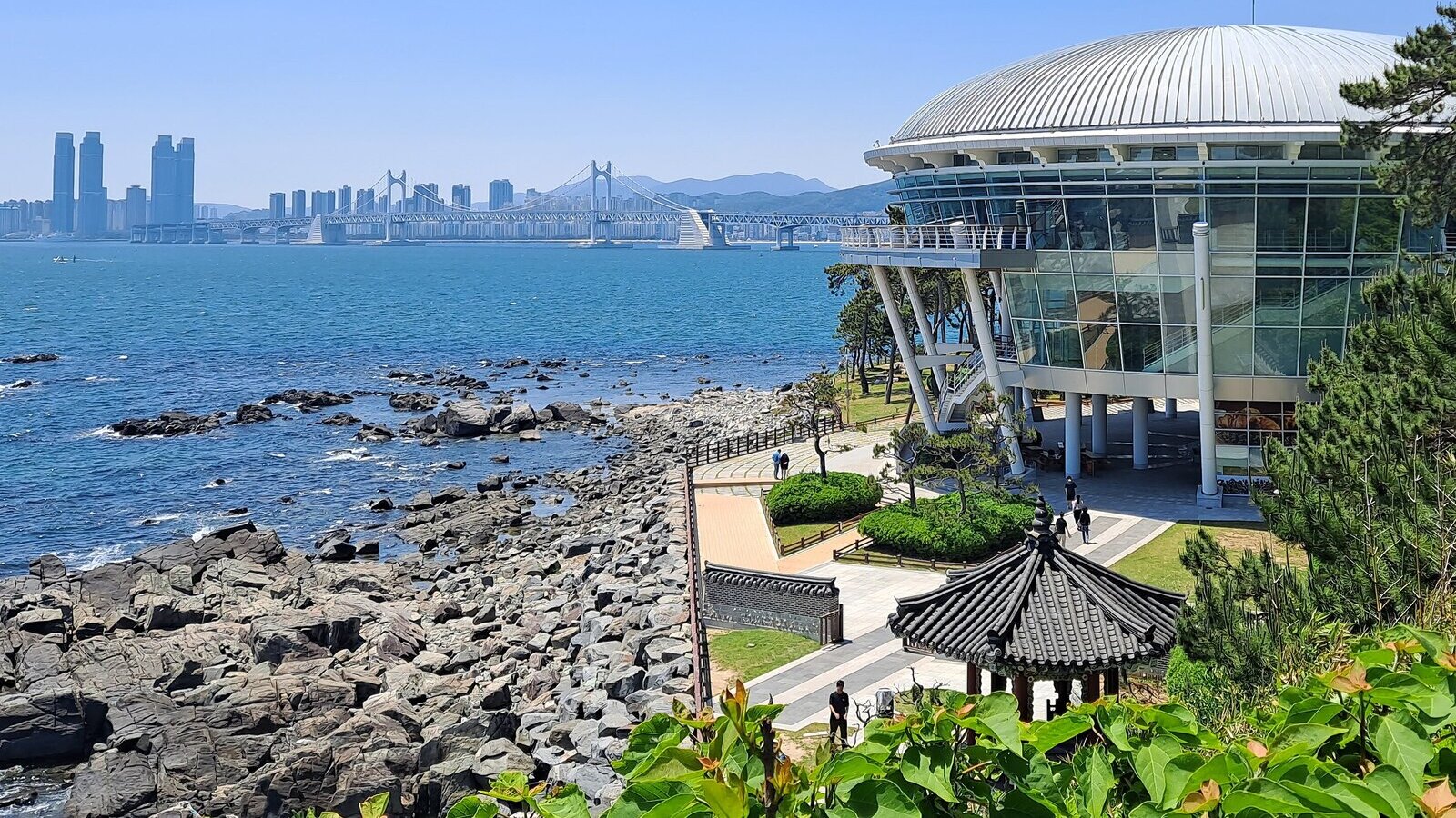 맑은 낮에 동백섬 언덕에서 본 풍경. 오른쪽에 둥근 지붕의 누리마루 APEC하우스와 기와 정자가 있고, 왼쪽 바위 해안 너머 바다 건너 멀리 광안대교와 고층 건물들이 보인다