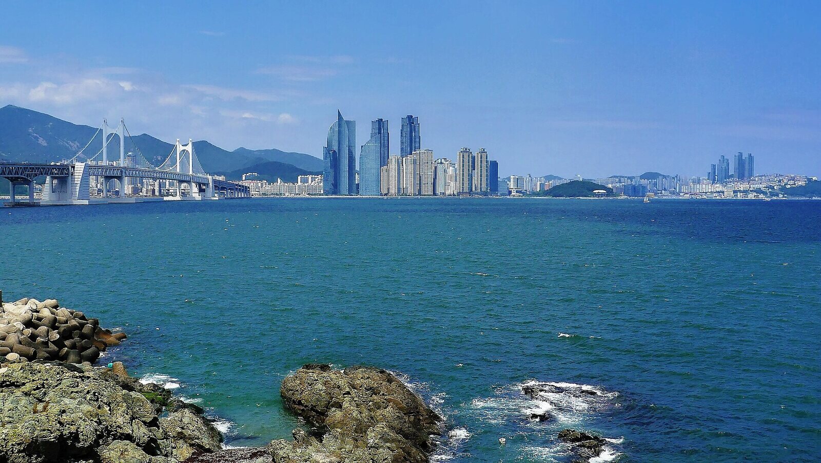 맑은 낮에 이기대 바닷가 바위와 방파제 블록 너머로 본 바다. 왼쪽에 광안대교, 가운데에 마린시티 고층 건물, 오른쪽에 동백섬과 해운대가 보인다