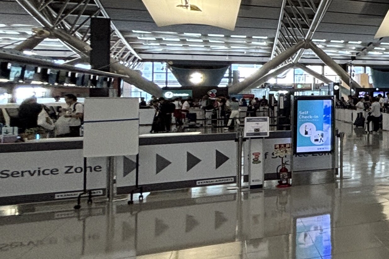 밝은 공항 출발 로비. 바닥에 불빛이 비치고, 화살표 무늬가 그려진 흰 칸막이 너머로 체크인 줄에 선 여행객들과 셀프 체크인 안내 화면이 보인다